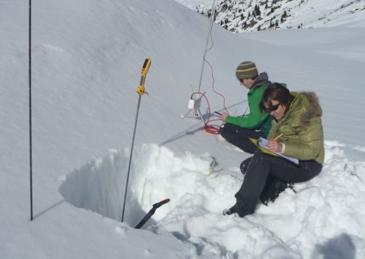 Foto mit 2 Studien-Mitarbeiter:innen, die bei einem Feldtest im tief verschneiten Gelände verschiedene Messroutinen zur Bestimmung der Schneehärte erheben