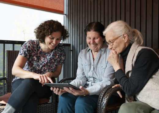 Drei Frauen vor einem Tablet, von denen eine digitale Unterstützungsmöglichkeiten für die Pflege zu Hause erläutert.